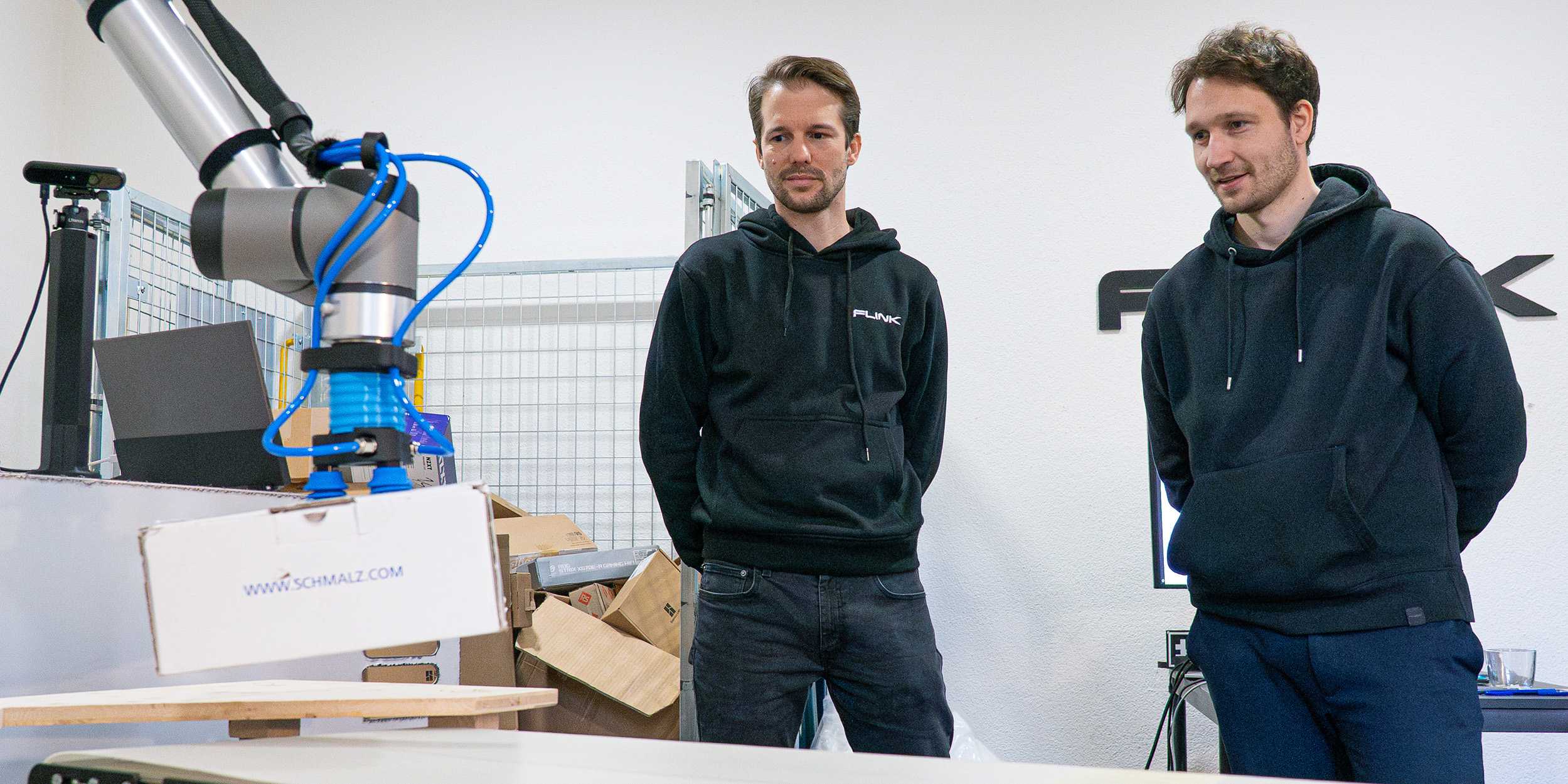 Moritz Geilinger und Simon Huber stehen neben einem Roboter, welcher ein Paket hochhebt