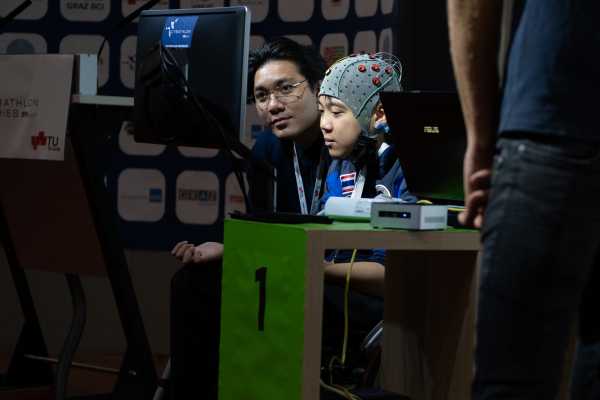 Two people sit in front of a computer screen, the person on the right wears a cap with red dots.