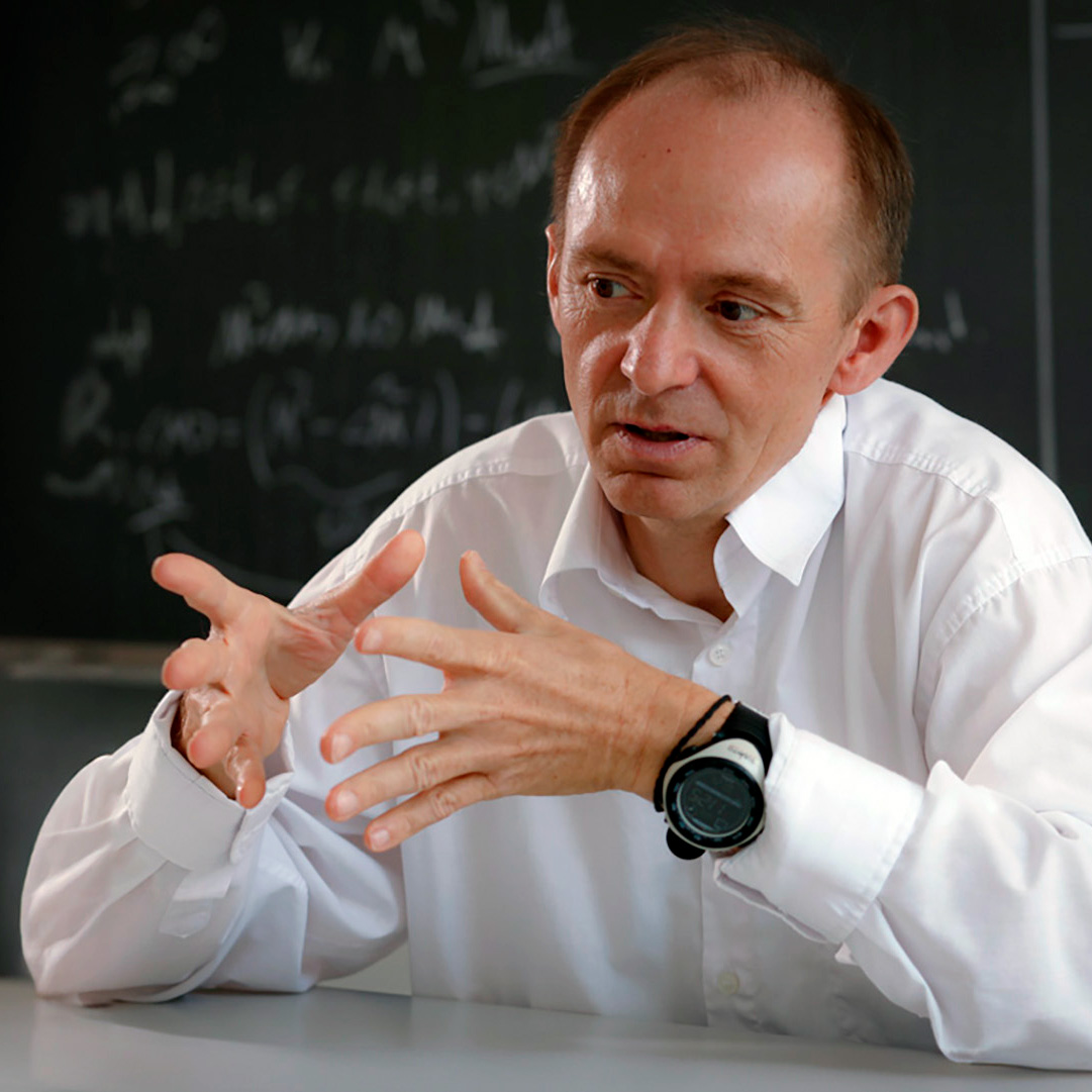Juraj Hromkovic gesturing and sitting in front of a blackboard.